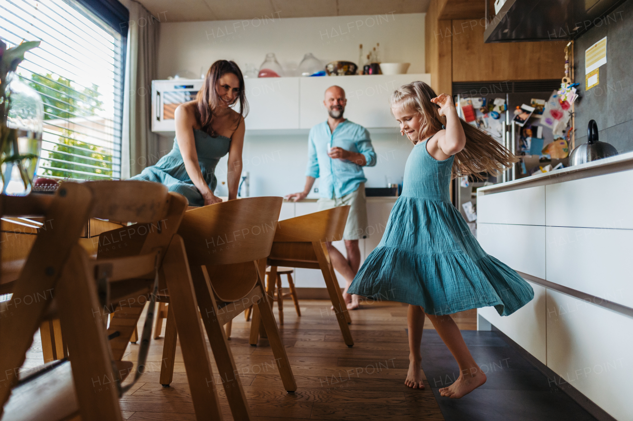 Little girl dancing in the kitchen, while parents watching her. Cheerful family having fun together in kitchen on sunny morning. Concept of family bonding.