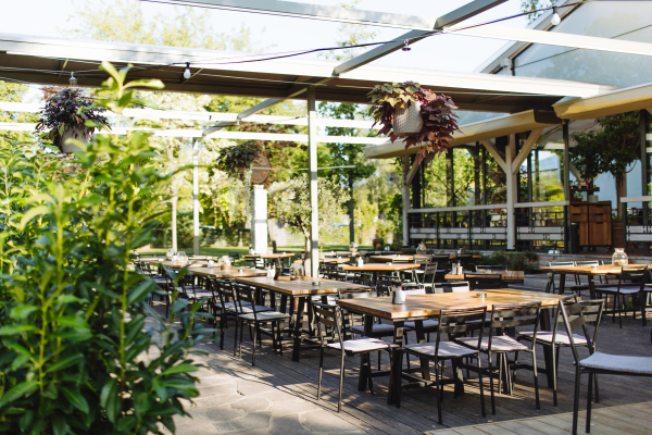 Covered summer terrace of a restaurant with wooden tables, chairs, and flooring. Restaurant patio made of natural materials with lot of greenery.