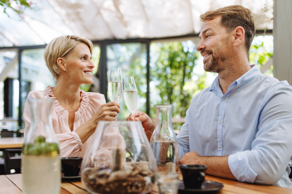 Portrait of beautiful couple in a restaurant, on a romantic date. Husband and wife are clinking champagne glasses, making a toast at restaurant patio.