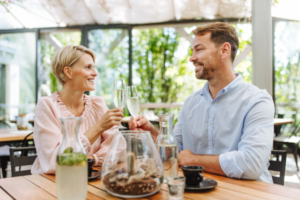 Portrait of beautiful couple in a restaurant, on a romantic date. Husband and wife are clinking champagne glasses, making a toast at restaurant patio.