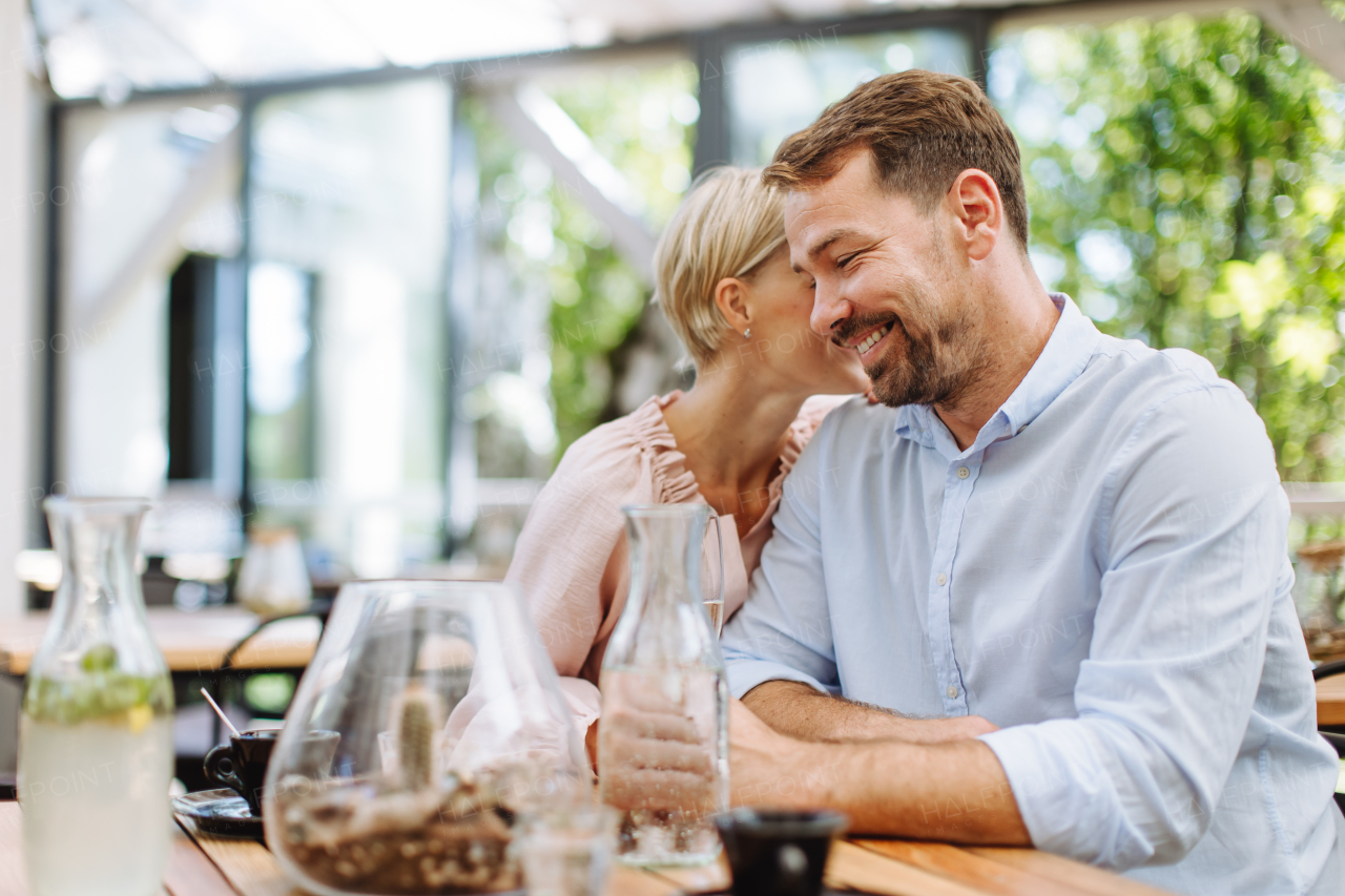 Portrait of beautiful couple in a restaurant, on a romantic date. Wife whispers into his husband's ear, having a romantic moment at restaurant patio.