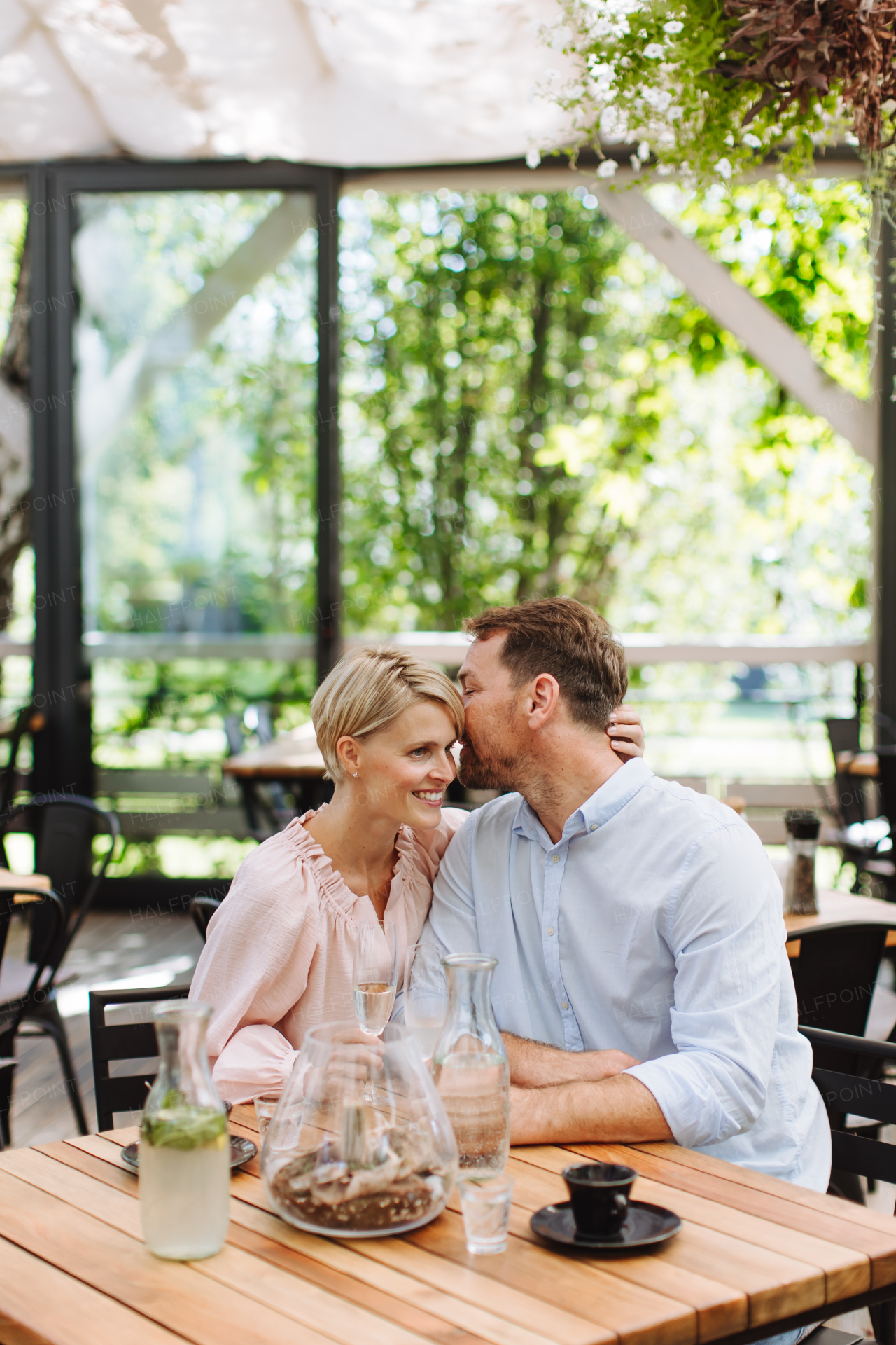 Portrait of beautiful couple in a restaurant, on a romantic date. Husband whispers into his wife's ear, having a romantic moment at restaurant patio.