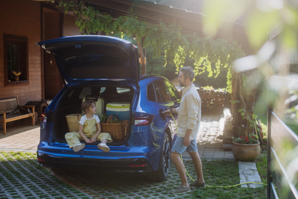 Father charging their electric car, little daughter sitting and waiting in car trunk.