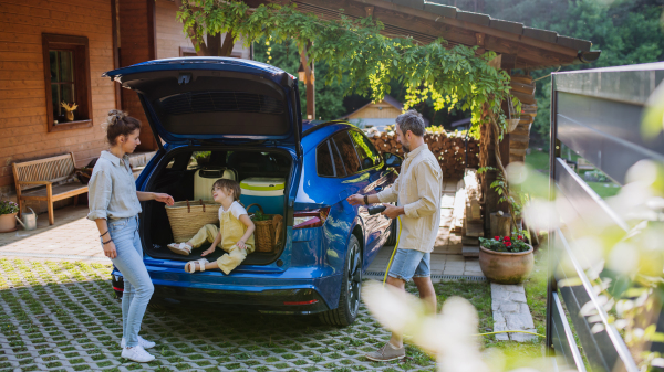 Father charging their electric car, little daughter and mother sitting and waiting in car trunk.