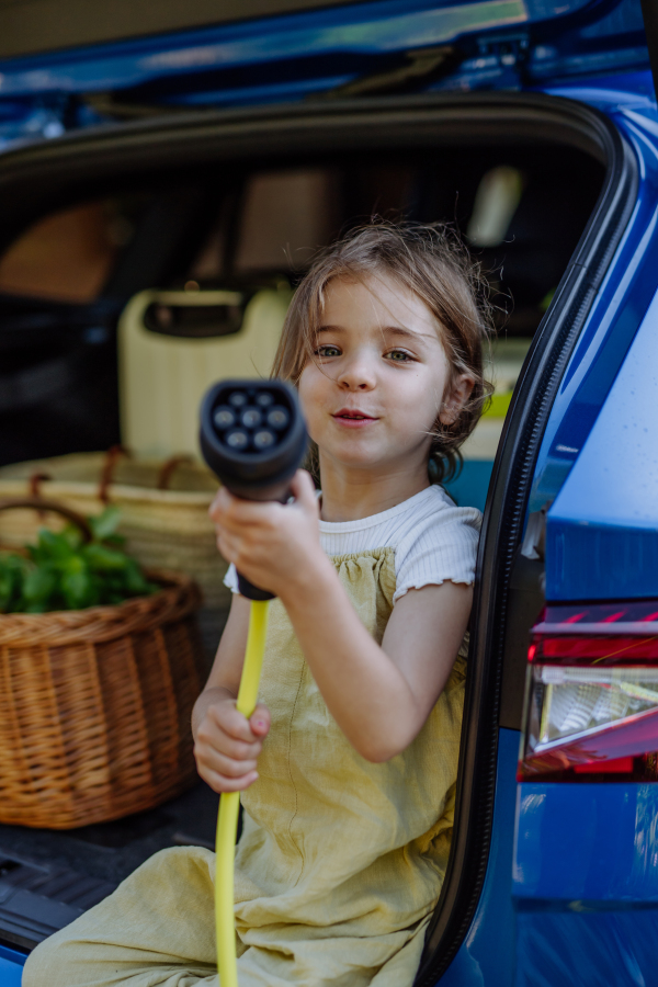 Little girl holding power supply cable, sitting in car trunk.