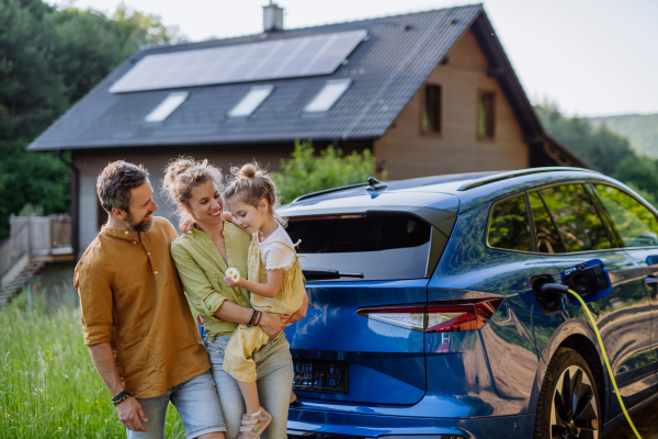 Family with little daughter standing in front of their house with solar panels on the roof, having electric car.