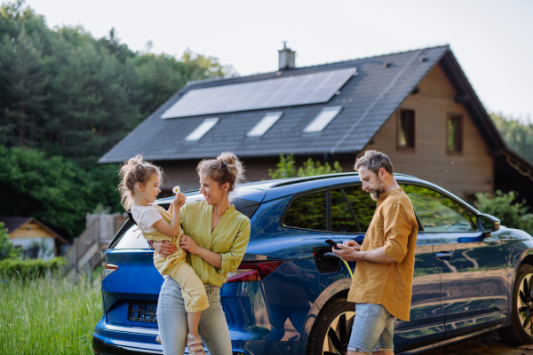 Family with little daughter standing in front of their house with solar panels on the roof, having electric car.