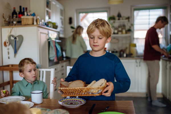 Family preparing breakfast in home kitchen in the morning. Oldest son bringing bread on the table.