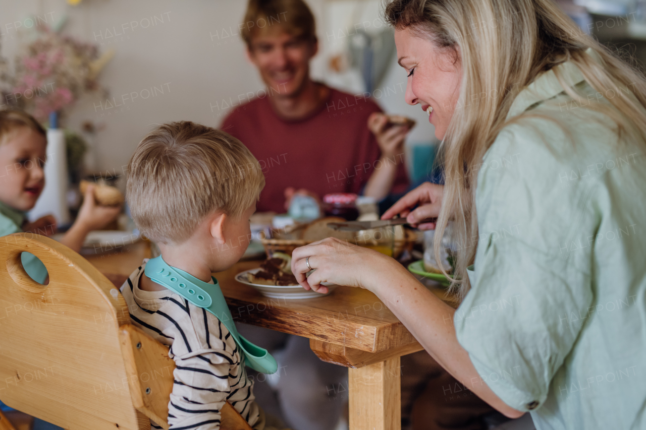 Young family eating breakfast together in home kitchen. Healty breakfast or snack before school and work. Mother helping youngest son.