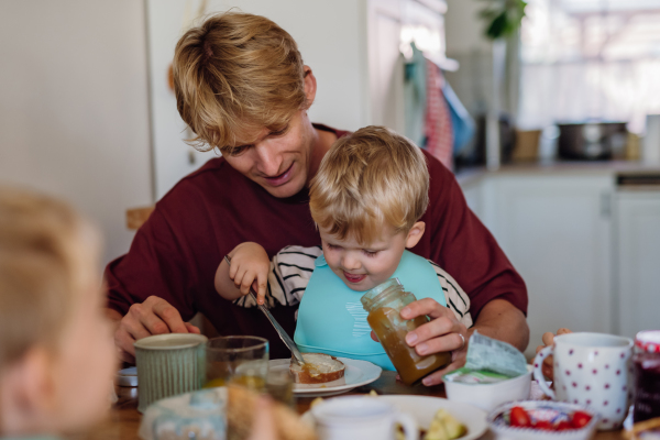 Father feeding cute little baby with breakfast in home kitchen. Boy spreading jam on his bread. Healthy breakfast or snack before day care.