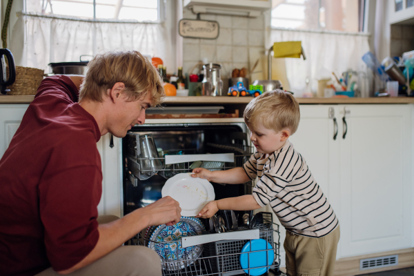 Little boy helping father to load dishwasher after together breakfast. Cleaning the kitchen before leaving to work and daycare. Family morning routine.