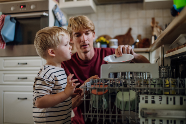 Little boy helping father to load dishwasher after together breakfast. Cleaning the kitchen before leaving to work and daycare. Family morning routine.