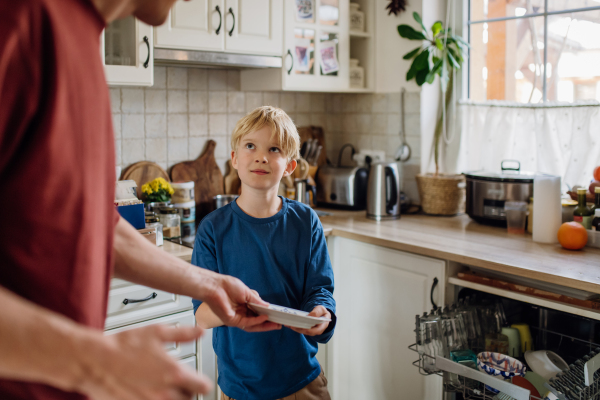 Young son helping father to load dishwasher after breakfast. Cleaning the kitchen before leaving to work and school. Family morning routine.