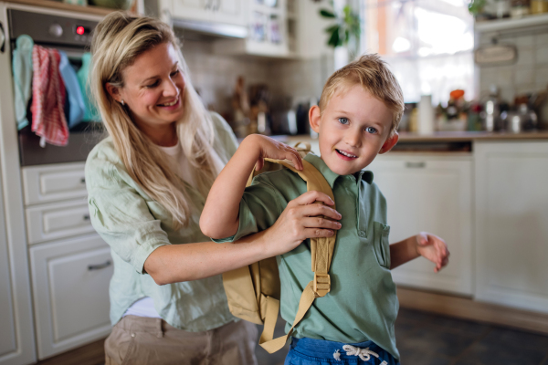 Mother saying goodbye to son before school, helping him to put backpack on his back in kitchen.