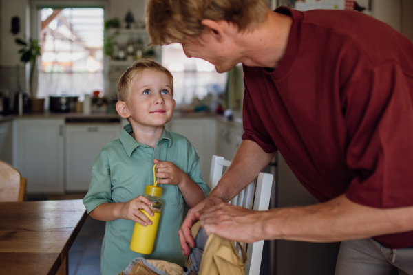 Dad is packing snack for his son for school. Putting bottle with juice and lunch box with healthy snack into his school bag for lunch.