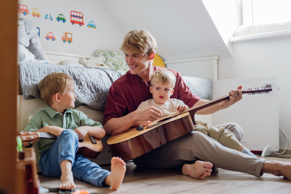 Father teaching boys to play on guitar. Boys having fun in their room with dad, playing guitar and singing. Concept of Father's Day, and fatherly love.