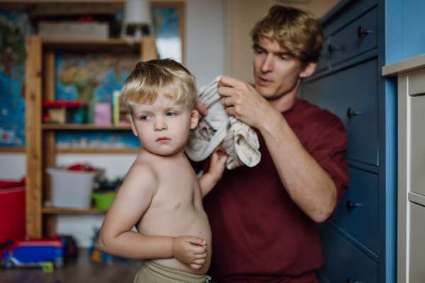 Father putting shirt on little son, changing from pajamas in the morning. Boy is sad, crying. Concept of defiant toddler.