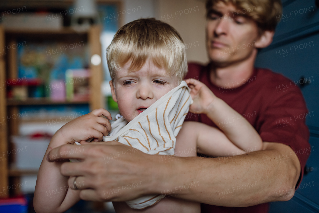 Father putting shirt on fussy little son, changing from pajamas in the morning. Boy is sad, crying. Concept of defiant toddler.