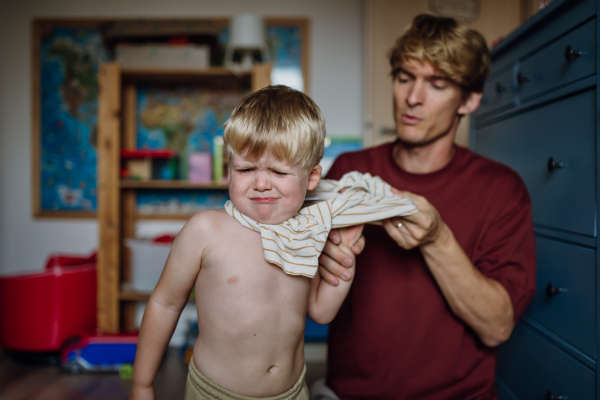 Father putting shirt on little son, changing from pajamas in the morning. Boy is sad, crying. Concept of defiant toddler.