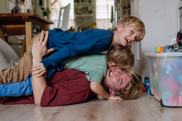 Portrait of father embracing his two sons at home, having fun, lying on floor and laughing. Dad and boys spending quality time together, bonding.