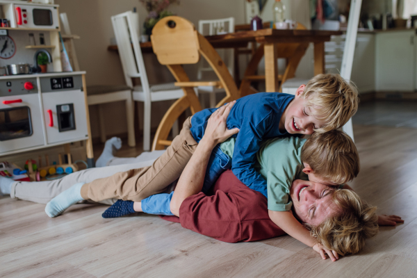 Portrait of father embracing his two sons at home, having fun, lying on floor and laughing. Dad and boys spending quality time together, bonding.