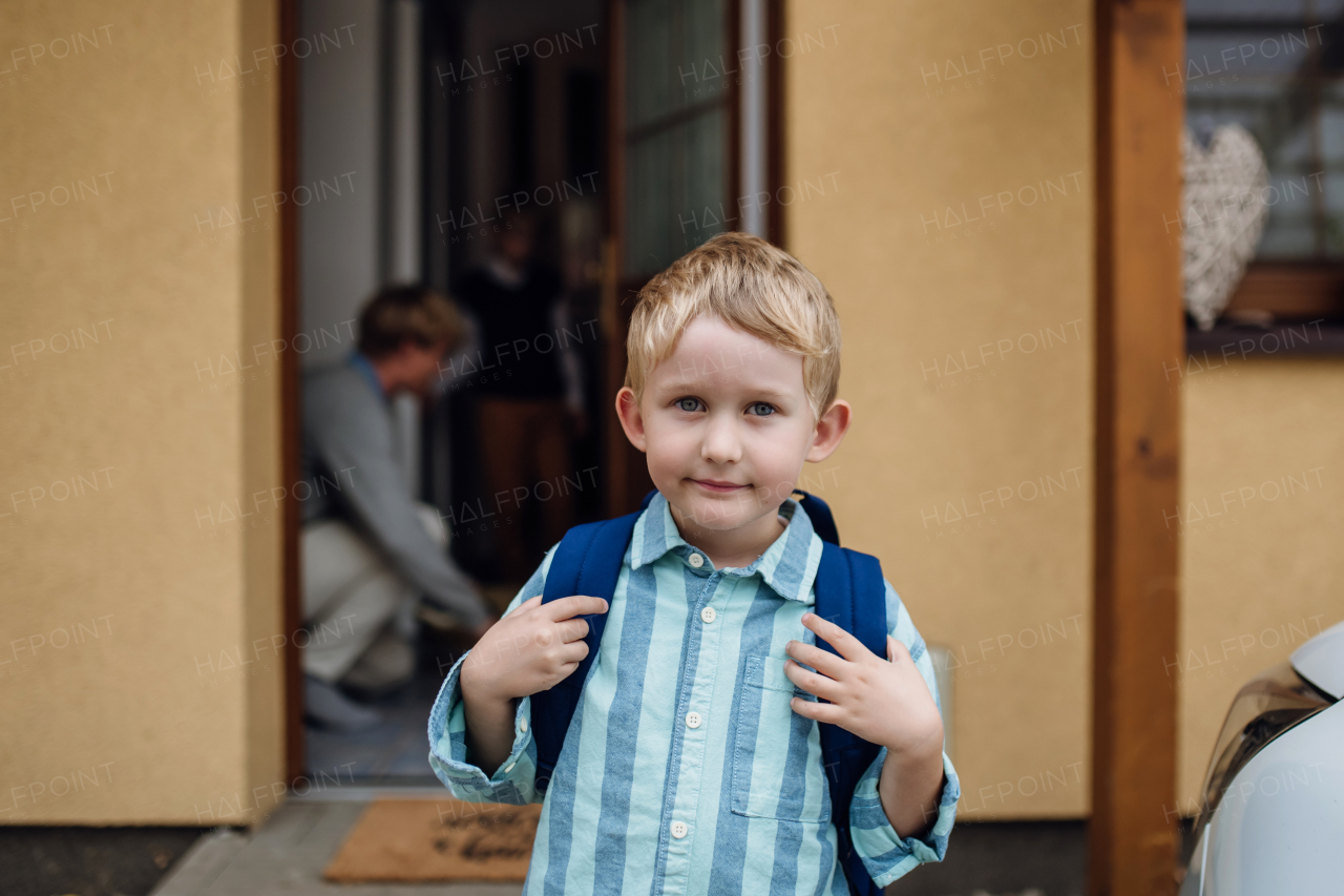 Portrait of schoolboy with backpack on back standing in front of house. Preparation for school day, in the morning.