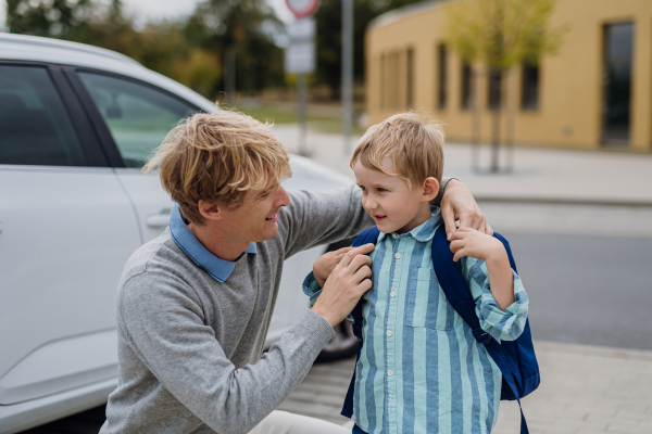 Father saying goodbyeto to son in front of school building, hugging him and helping him with backpack. Dad heading to work. Concept of work-life balance for parents.