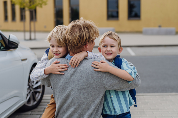 Father saying goodbyeto to sons in front of school building, hugging both boys. Dad heading to work. Concept of work-life balance for parents.