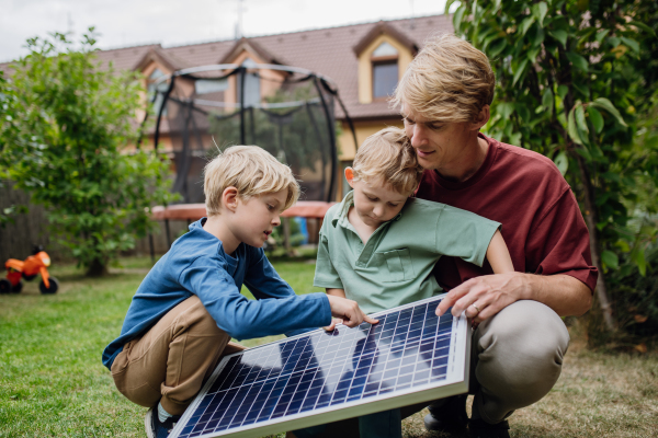 Father and his sons holding solar panel in the garden, learning about photovoltaics and green energy. Concept of sustainable living.