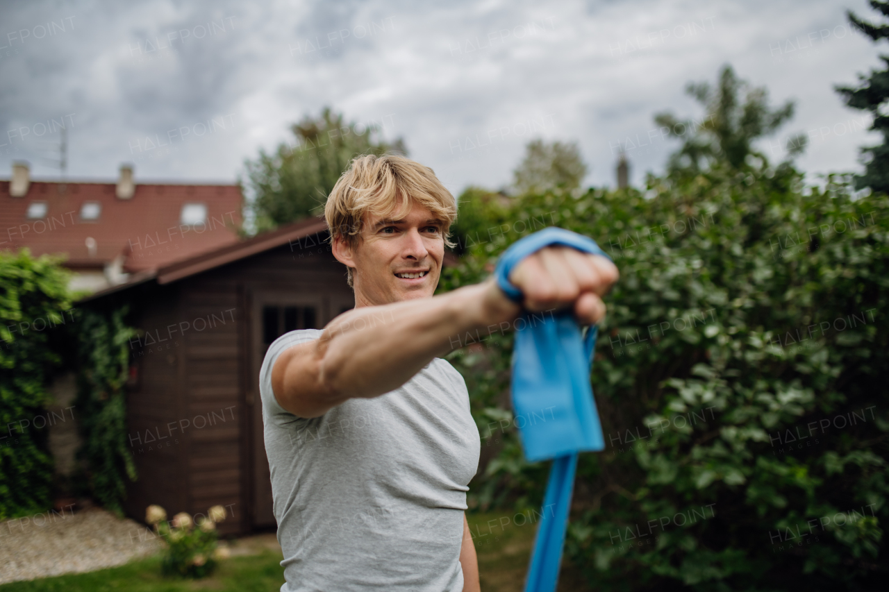 Middle aged man doing strength exercises with resistance band outdoors in the garden. Concept of workout routine at home.