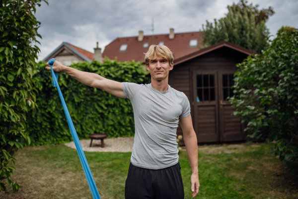 Middle aged man doing strength exercises with resistance band outdoors in the garden. Concept of workout routine at home.