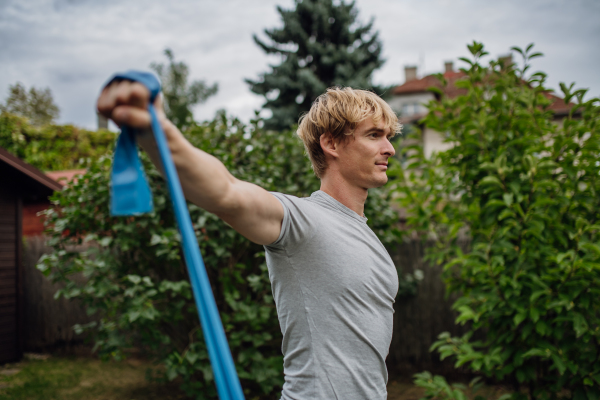 Middle aged man doing strength exercises with resistance band outdoors in the garden. Concept of workout routine at home.