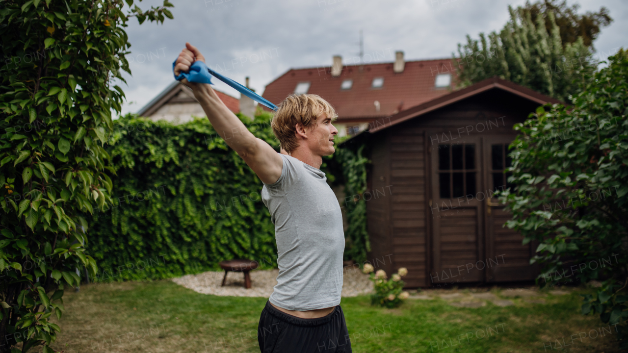 Middle aged man doing strength exercises with resistance band outdoors in the garden. Concept of workout routine at home.