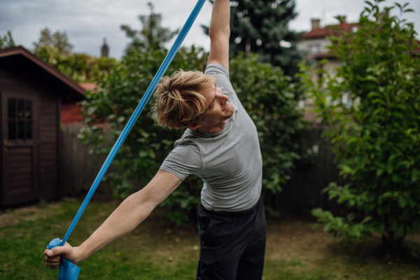 Middle aged man doing strength exercises with resistance band outdoors in the garden. Concept of workout routine at home.