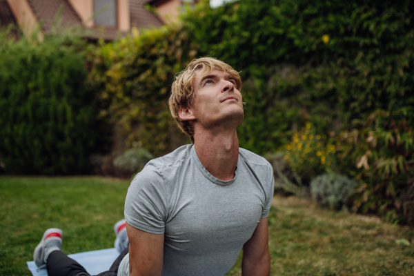 Middle aged man stretching, exercising outdoors in garden, doing yoga, pilates on the gym mat. Concept of workout routine at home.