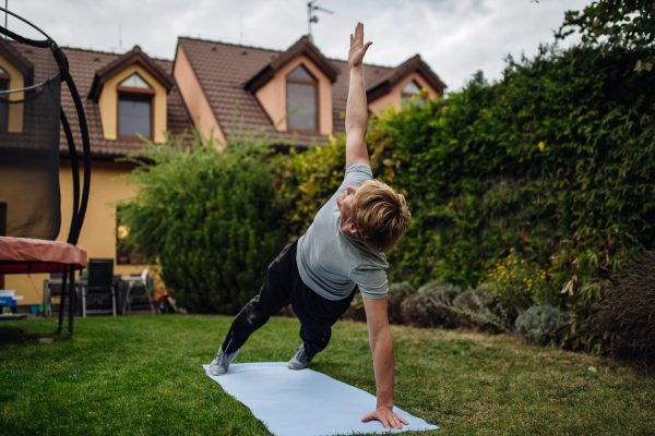 Middle aged man stretching, exercising outdoors in garden, doing yoga, pilates on the gym mat. Concept of workout routine at home.