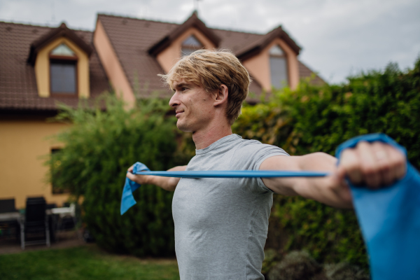 Middle aged man doing strength exercises with resistance band outdoors in the garden. Concept of workout routine at home.