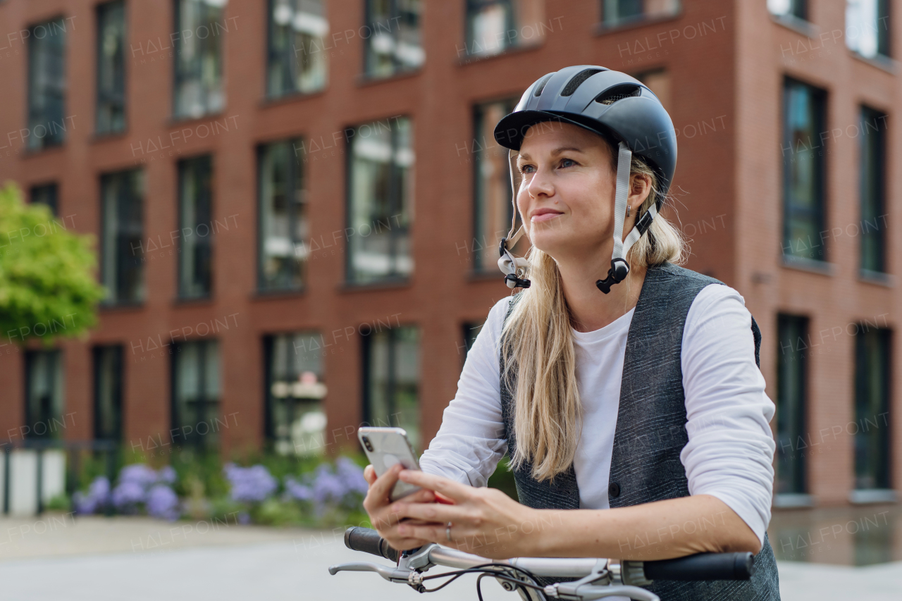 Beautiful middle-aged woman commuting through the city by bike, holding smartphone. Female city commuter traveling from work by bike after a long workday.