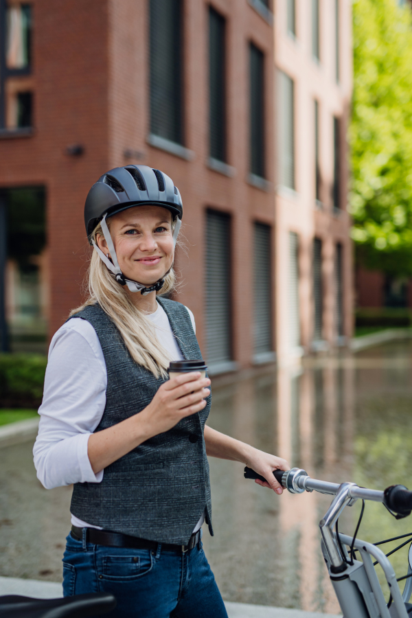 Beautiful middle-aged woman commuting through the city, buying, drinking coffe in front of office. Female city commuter with helmet traveling from work by bike after a long workday.