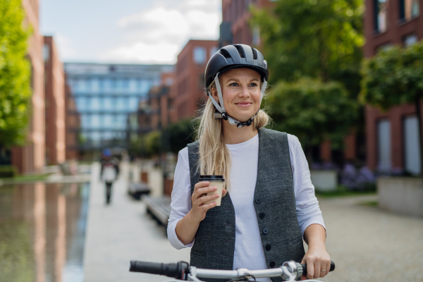 Beautiful middle-aged woman commuting through the city, buying, drinking coffe in front of office. Female city commuter with helmet traveling from work by bike after a long workday.