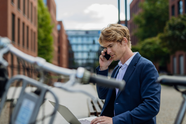 Portrait of businessman, freelancer or manager working outdoors in city park. Man with laptop on knees making call on smartphone, sitting on park bench during sunny day. Concept of working remotely.