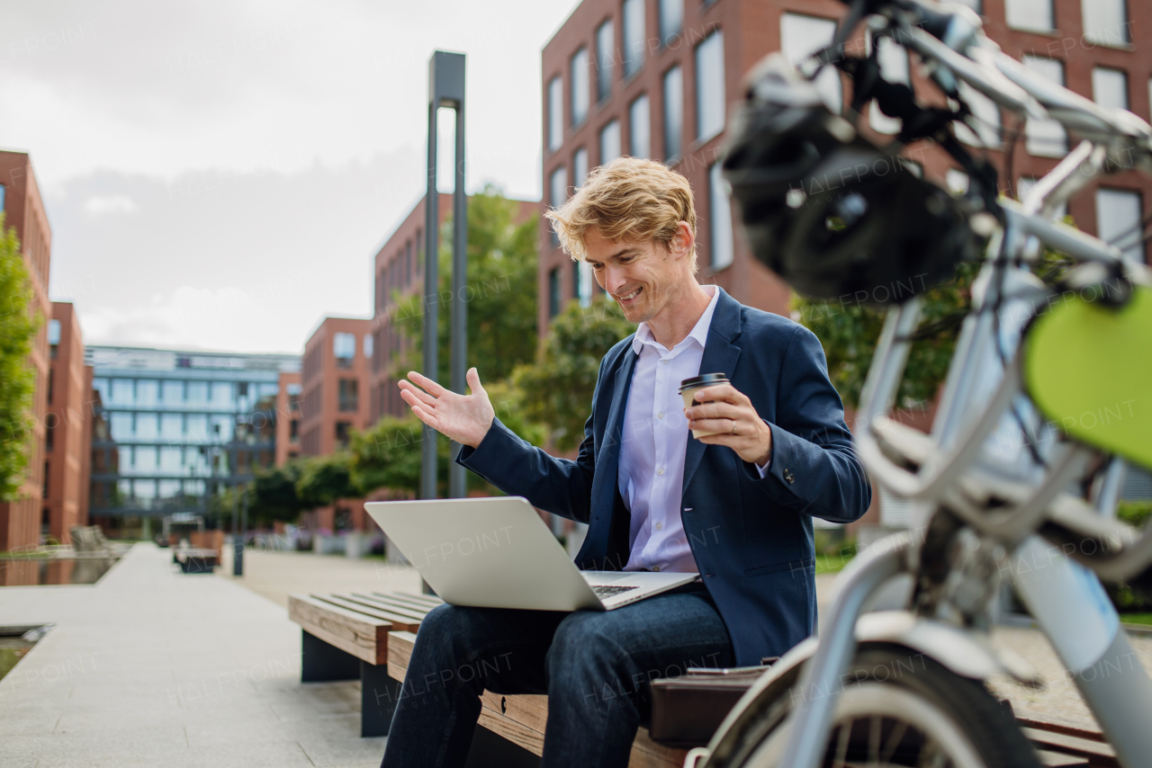 Portrait of businessman, freelancer or manager working outdoors in city park. Man with laptop on knees drinking coffee, having video call, listening music. Concept of working remotely.