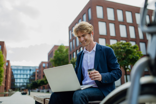 Portrait of businessman, freelancer or manager working outdoors in city park. Man with laptop on knees drinking coffee, having video call, listening music. Concept of working remotely.