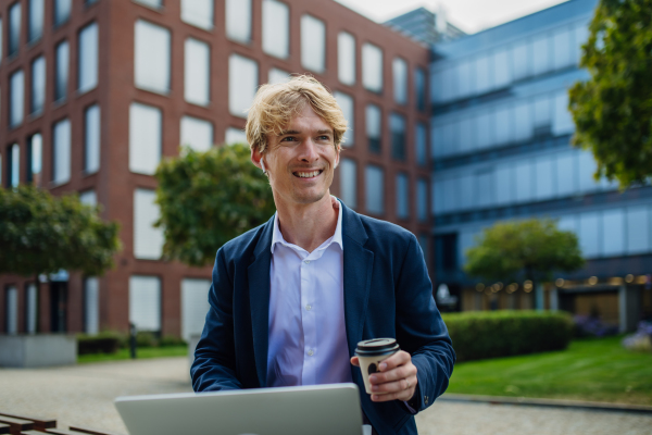 Portrait of businessman, freelancer or manager working outdoors in city park. Man with laptop on knees drinking coffee, having video call, listening music. Concept of working remotely.