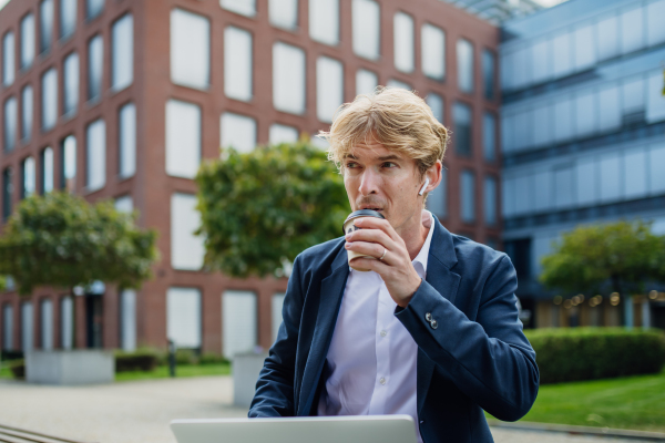 Portrait of businessman, freelancer or manager working outdoors in city park. Man with earbuds in ears, drinking coffee from travel mug, istening music. Concept of working remotely.