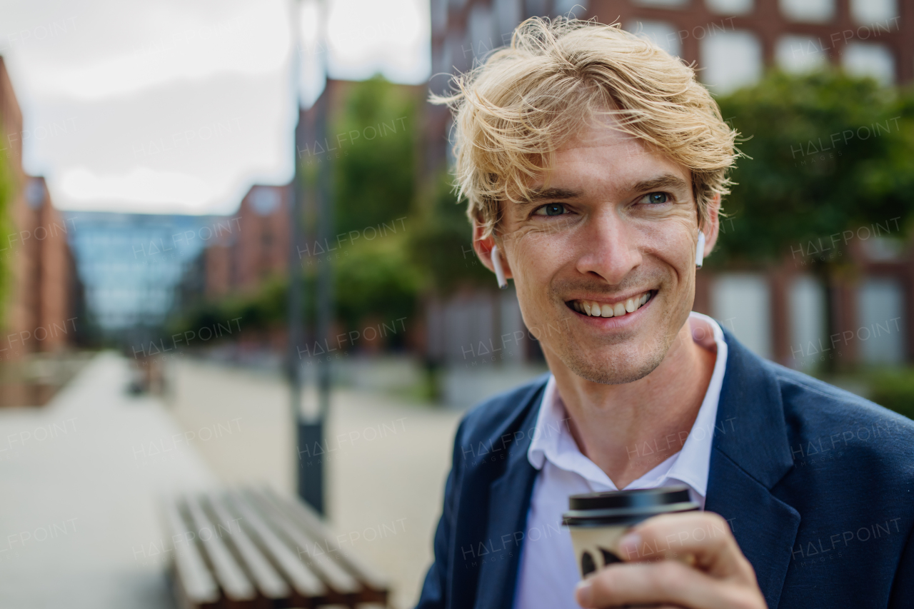 Portrait of businessman, freelancer or manager working outdoors in city park. Man with earbuds in ears, drinking coffee, having video call, listening music. Concept of working remotely.