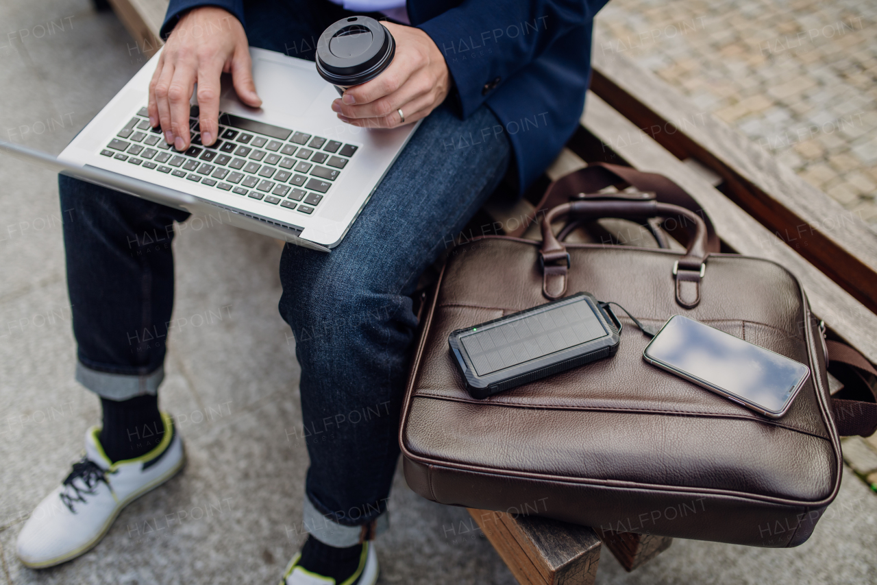 Portrait of businessman, freelancer or manager working outdoors in city park. Man with laptop on knees drinking coffee, charging his smartphone with solar phone charger. Concept of working remotely.