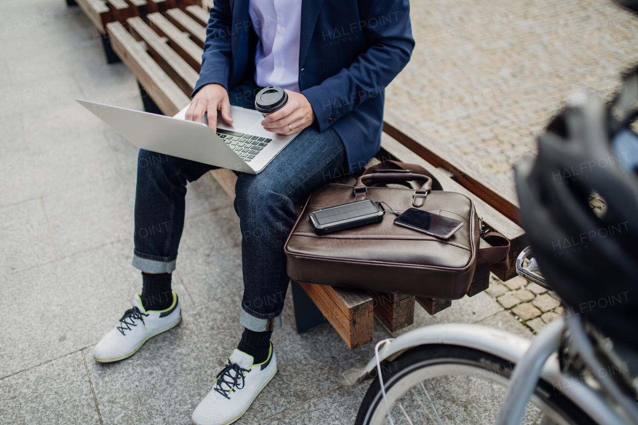 Portrait of businessman, freelancer or manager working outdoors in city park. Man with laptop on knees drinking coffee, charging his smartphone with solar phone charger. Concept of working remotely.