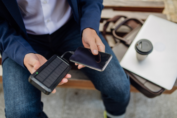 Portrait of businessman, freelancer or manager working outdoors in city park. Man with laptop on knees drinking coffee, charging his smartphone with solar phone charger. Concept of working remotely.