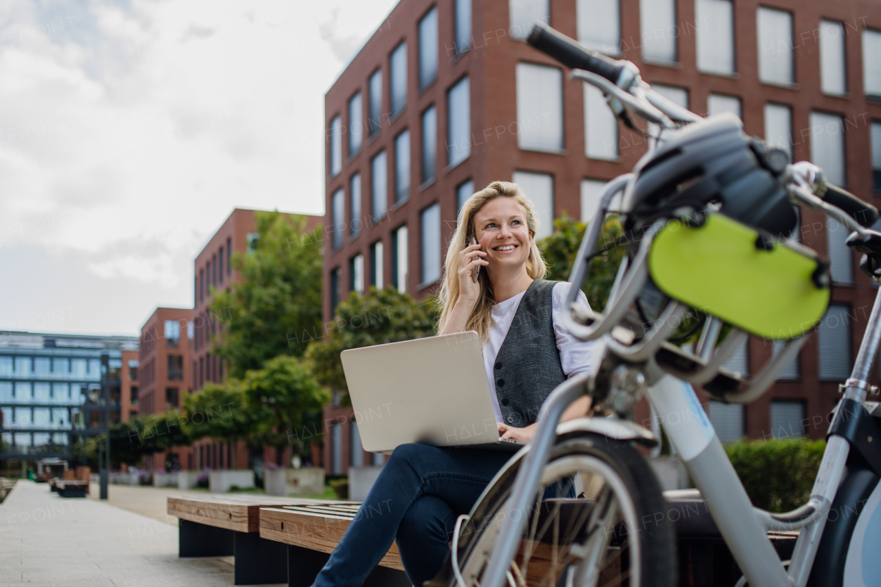 Portrait of businesswoman, freelancer or manager working outdoors in city park. Woman with laptop on knees making call on smartphone, sitting on park bench during sunny day. Concept of working remotely.